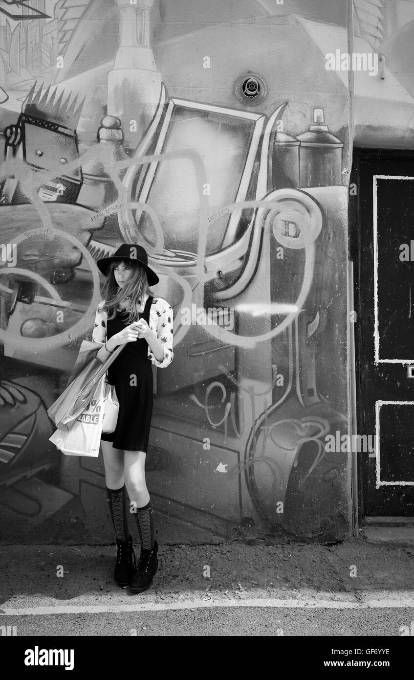Jeune fille de monochrome en jupe courte et hat en face de l'écriture graffiti holding shopping bags l'attente de quelqu'un, à la cool Banque D'Images