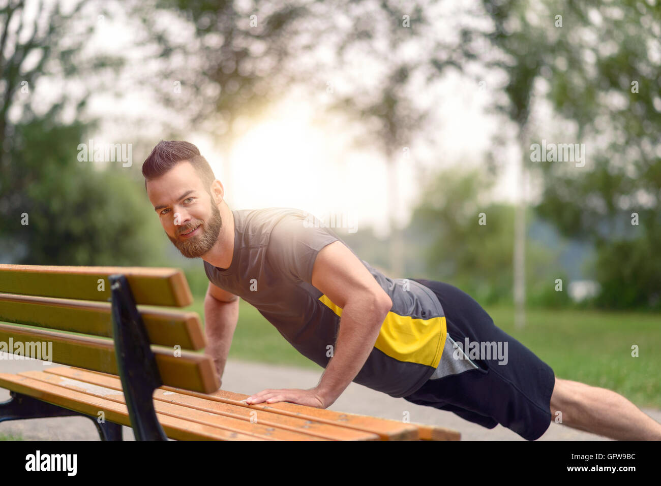 Jeune homme travaillant sur faire des tractions sur un banc de parc en bois comme il se réchauffe pour son entraînement quotidien ou de jogging Banque D'Images