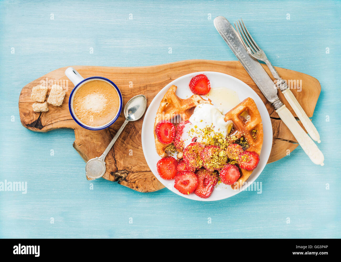 Le petit-déjeuner ensemble. Maison chaleureuse belgique gaufres à la crème fouettée, fraise, sirop d'érable et pistaches concassées, tasse de café, sucre brun olive sur planche de bois rustique sur fond peint bleu Banque D'Images