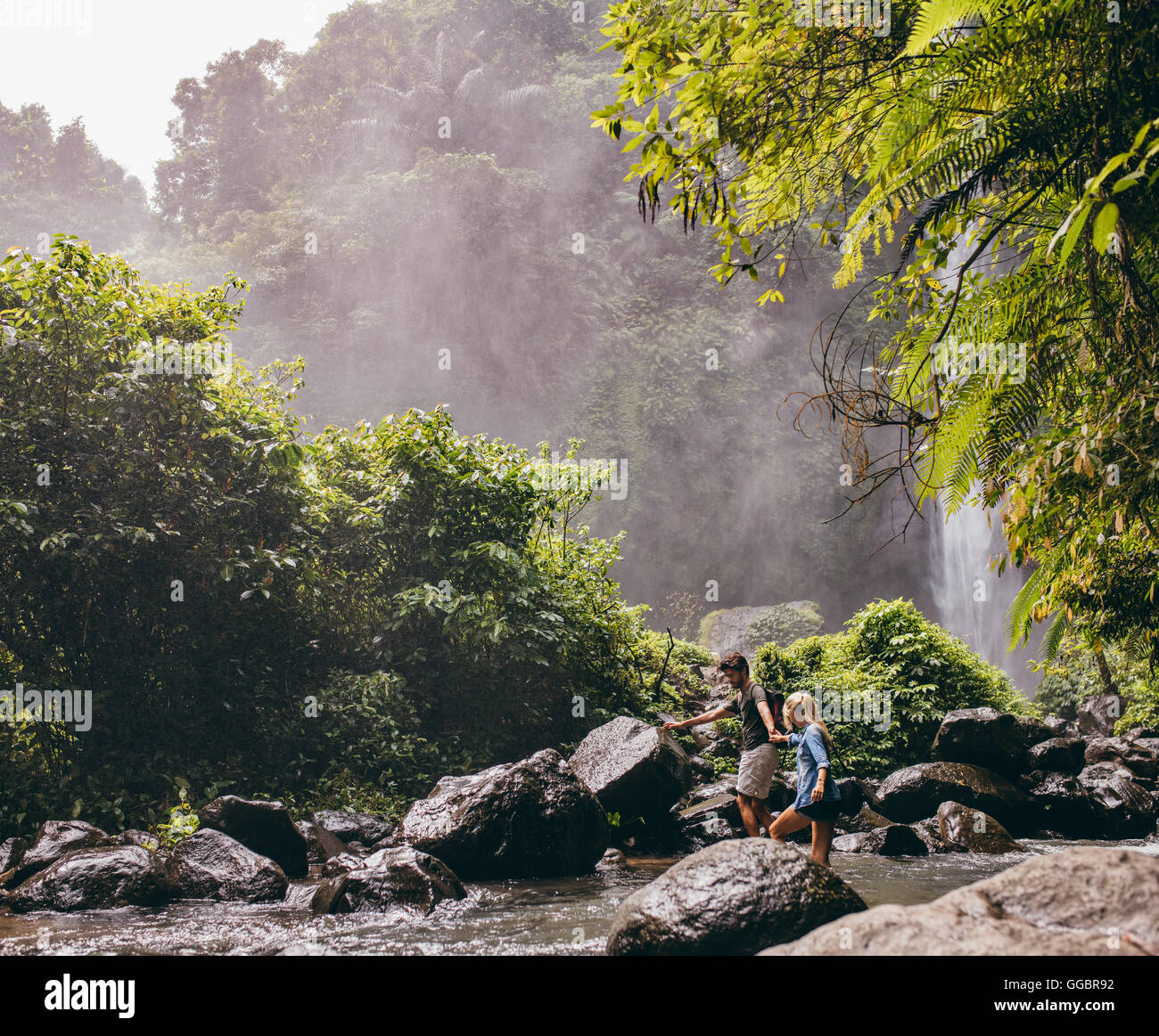 Couple traversant une rivière sur une randonnée dans la forêt. Jeune homme d'aider son amie en traversant un ruisseau. Banque D'Images