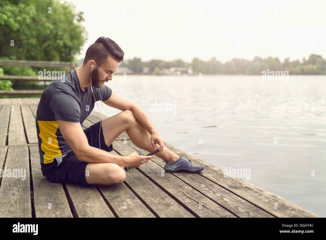 Beau jeune homme barbu de détente sur une terrasse en bois donnant sur un lac calme avec son téléphone portable dans sa main à la recherche Banque D'Images