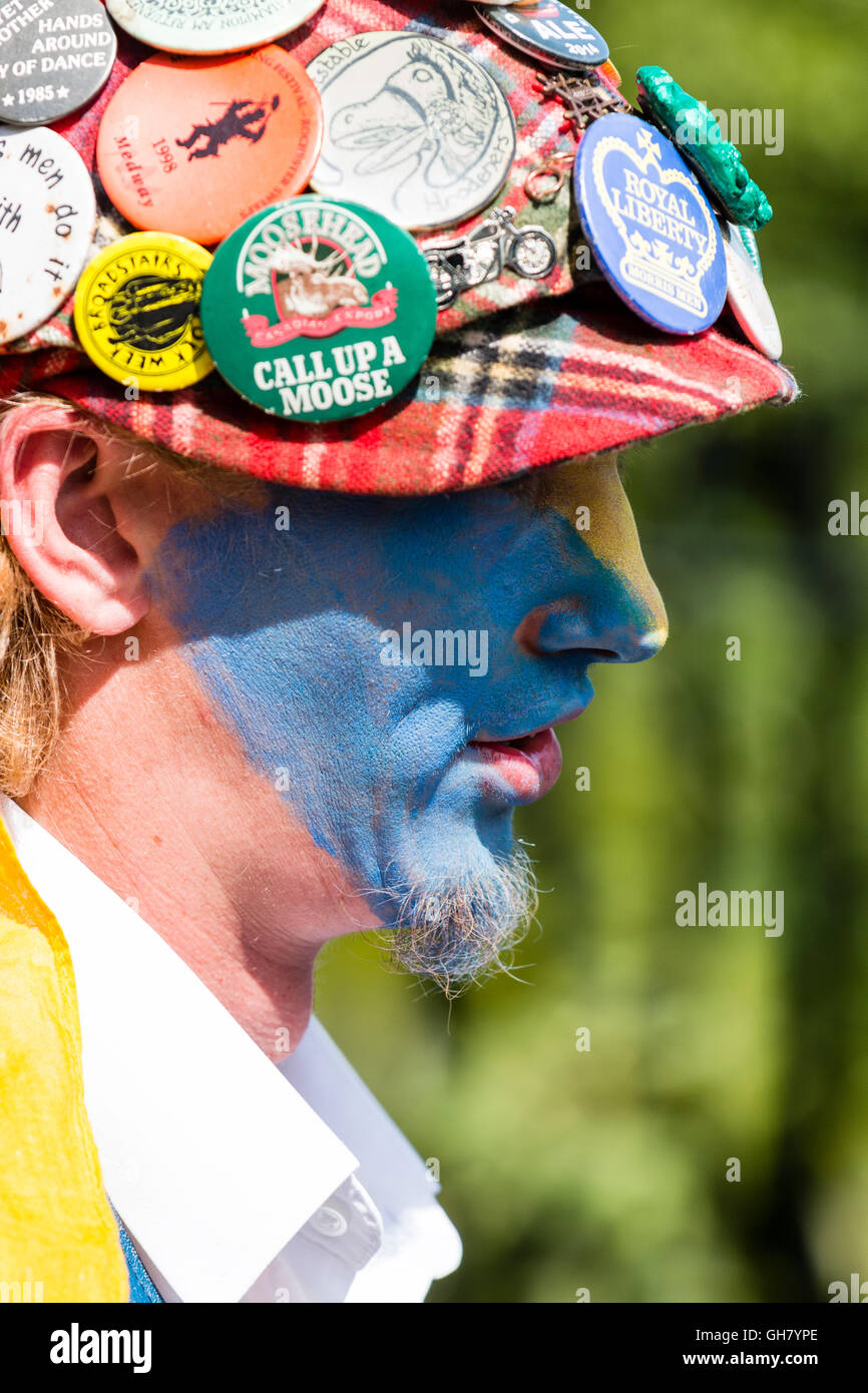 Close up of Royal Liberty Morris dancer, jeune homme, le visage peint en jaune et bleu, petite barbiche, porte la barbe bonnet rouge couvert de badges. Banque D'Images