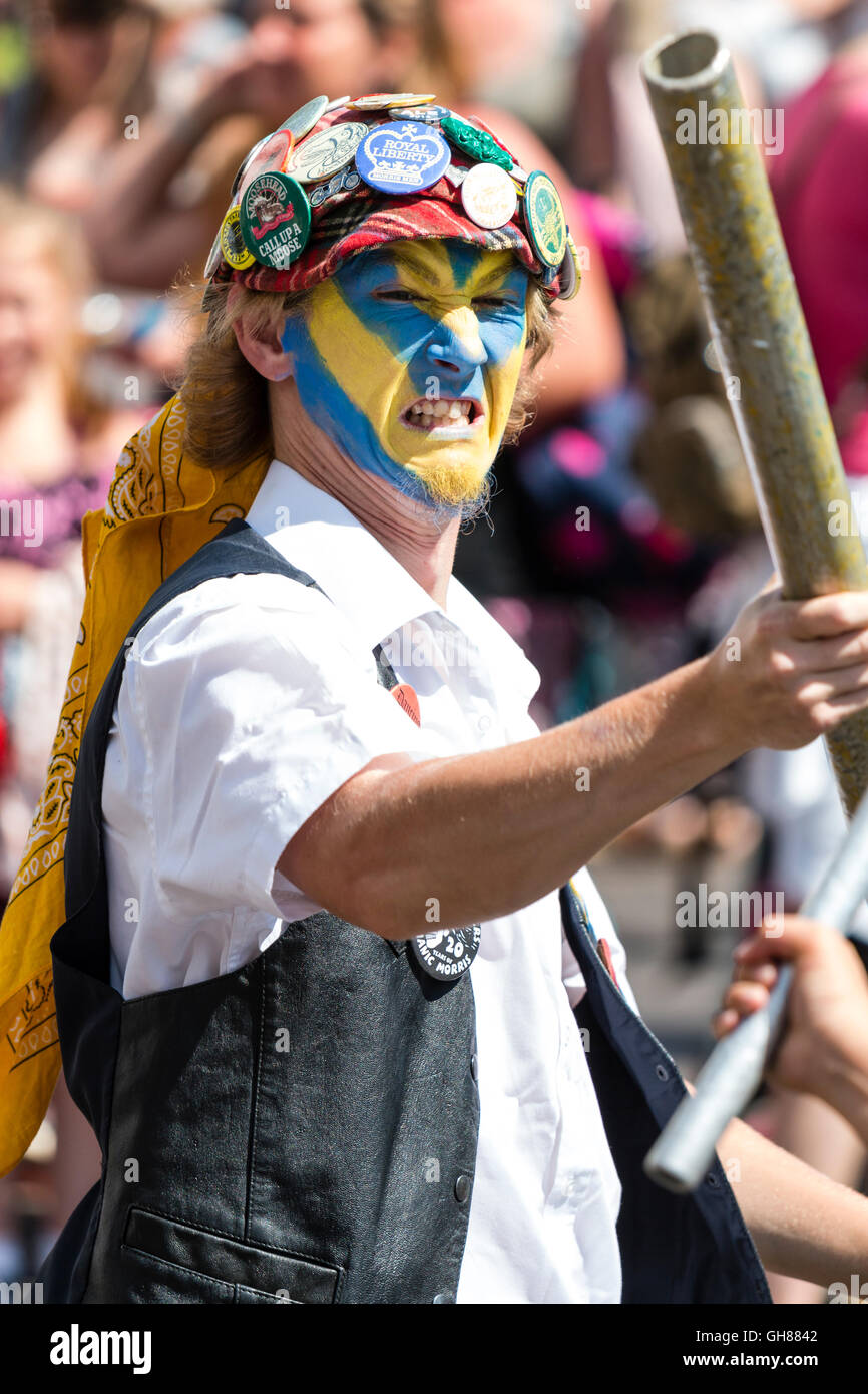 Close up of Royal Liberty Morris dancer, jeune homme, le visage peint en jaune et bleu, porte bouchon rouge couvert de badges, l'escrime avec poteau de métal. Banque D'Images