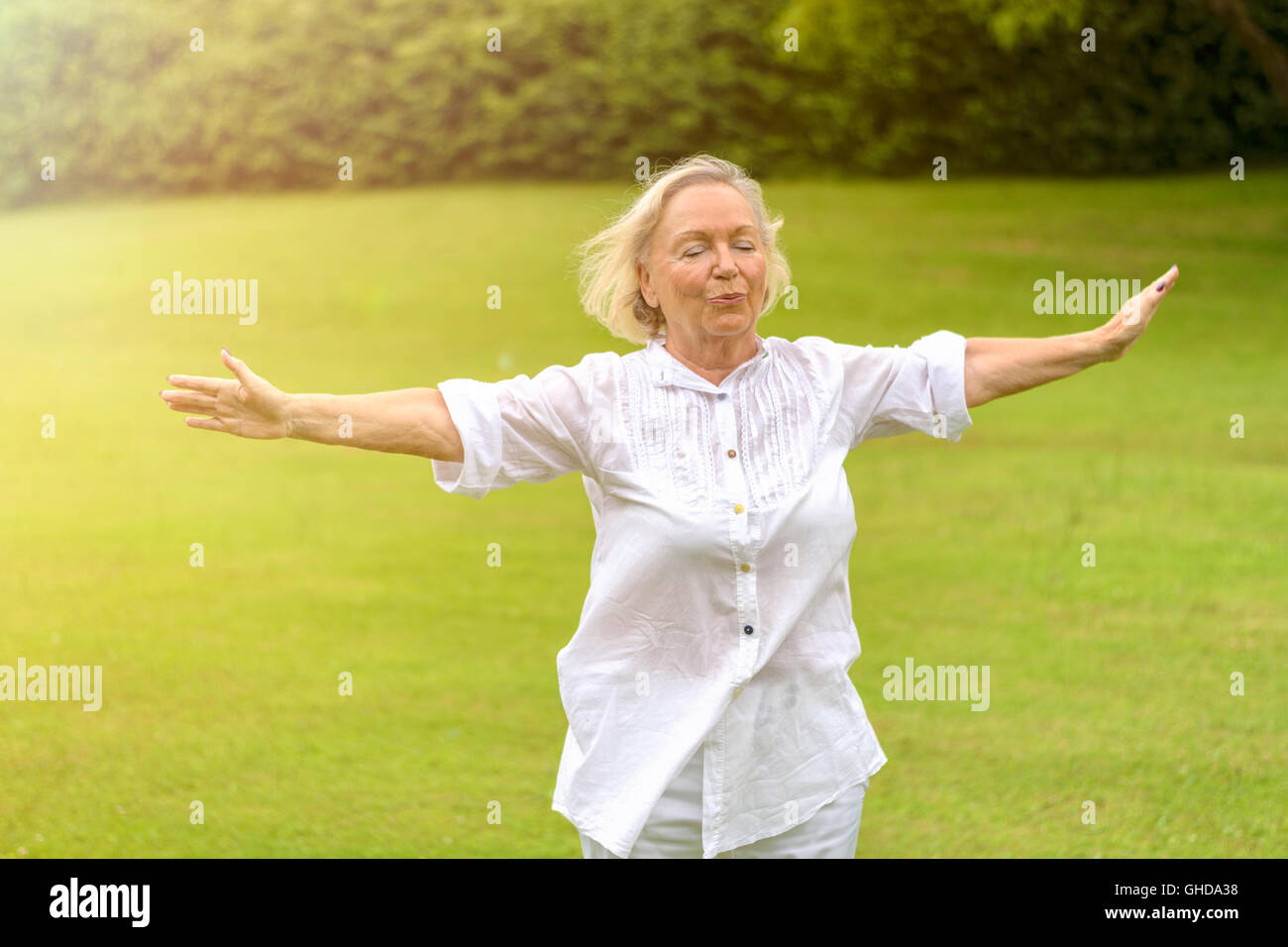 Seule belle senior woman in white rolled up long sleeve blouse de l'exercice à l'extérieur en fermant les yeux sur l'herbe verte avec l'exemplaire Banque D'Images