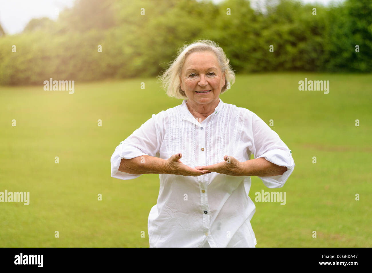 Seule belle senior woman in white rolled up long sleeve blouse pratiquer des exercices de respiration à l'extérieur sur l'herbe verte avec c Banque D'Images