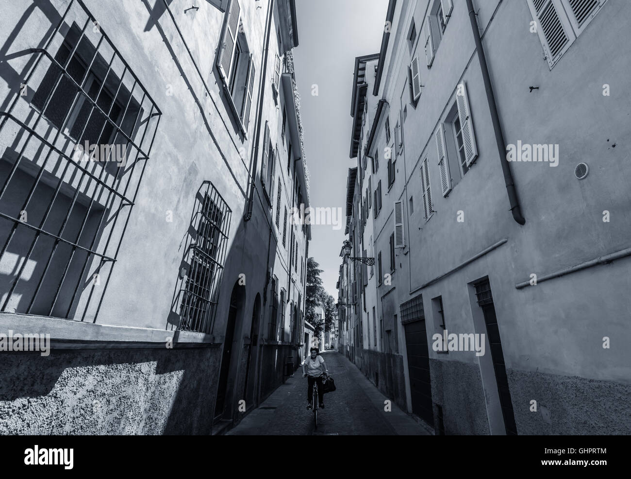 Une dame sur un vélo à vélo sur une route étroite entre deux bâtiments dans le centre de Parme, Italie, un matin de mai (noir et blanc) Banque D'Images