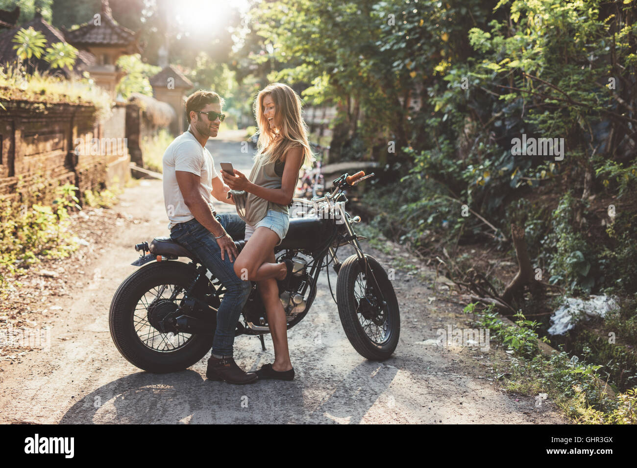 Tourné en plein air de cheerful young couple avec moto sur route de campagne. Homme assis sur moto avec femme tenant un smartphone. Banque D'Images