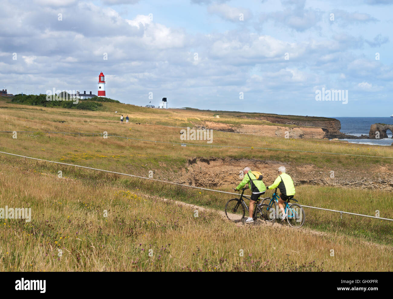 Vieux couple à vélo sur le sentier du littoral avec Souter Leuchtturm derrière, Whitburn, Tyne and Wear, England, UK Banque D'Images