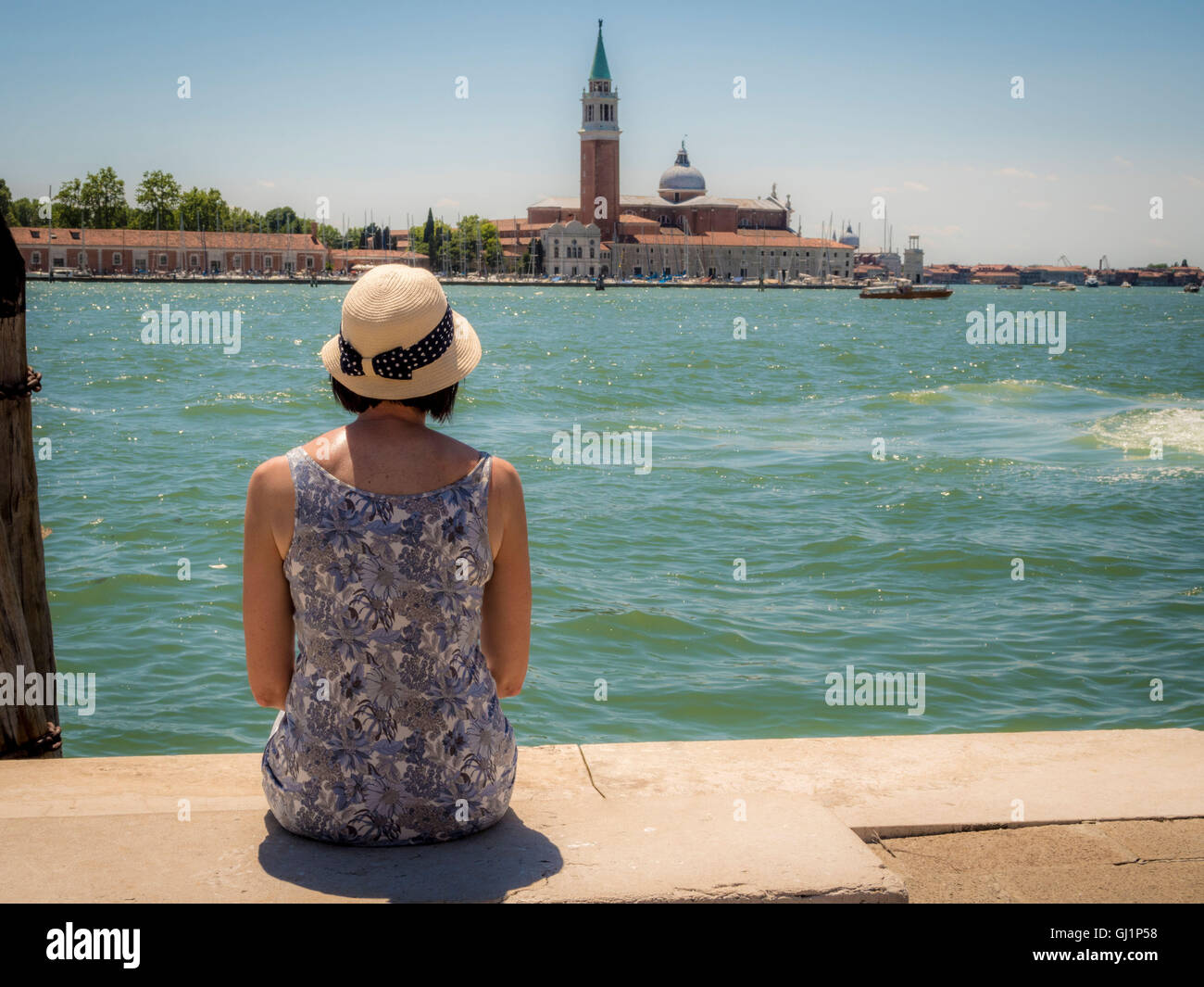 Vue arrière d'une femme, portant un chapeau de paille, assise sur un banc à l'ensemble de l'église et de l'île de San Giorgio Maggi Banque D'Images