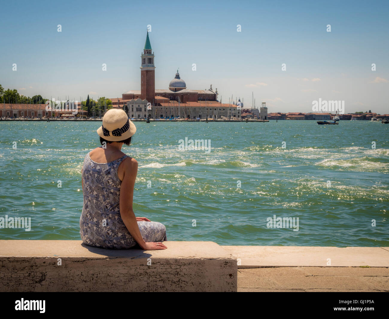Vue arrière de femme, portant un chapeau de paille, assise sur un banc à l'ensemble de l'église et de l'île de San Giorgio Maggiore. Banque D'Images