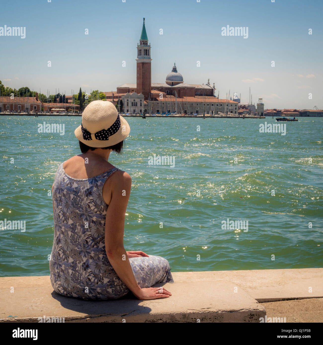 Vue arrière d'une femme, portant un chapeau de paille, assise sur un banc à l'ensemble de l'église et de l'île de San Giorgio Maggi Banque D'Images