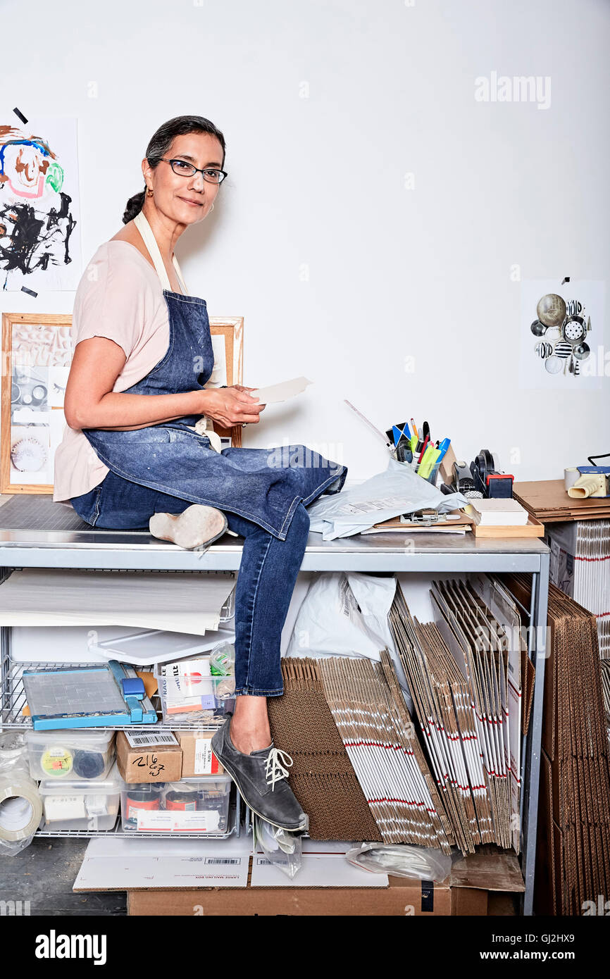 Woman sitting on desk portant un tablier looking at camera smiling Banque D'Images