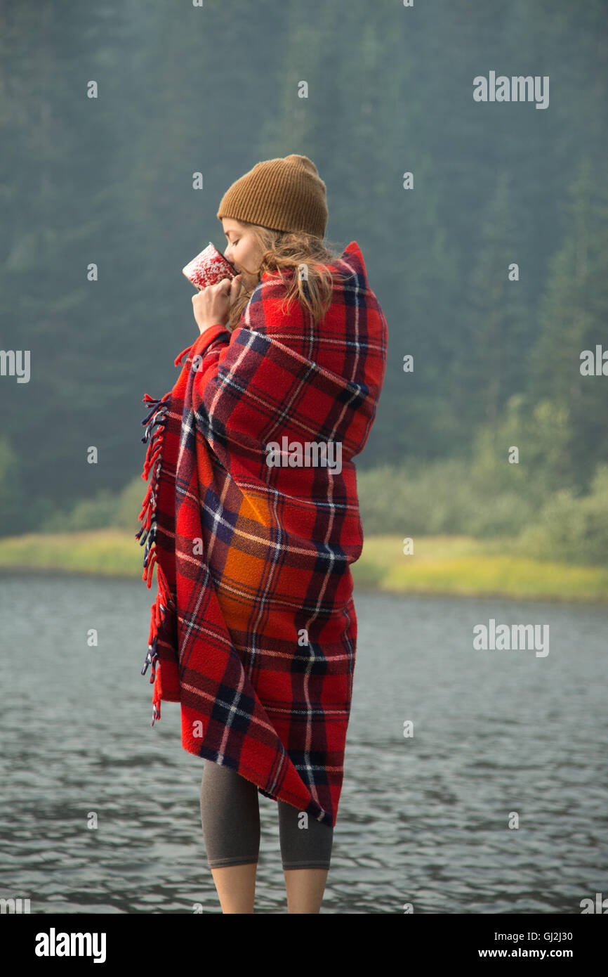 Femme enveloppée dans une couverture tartan boire du café par le lac, Mount Hood National Forest, North Carolina, USA Banque D'Images Femme enveloppée dans une couverture tartan boire du café par le lac, Mount Hood National Forest, North Carolina, USA Banque D'Images