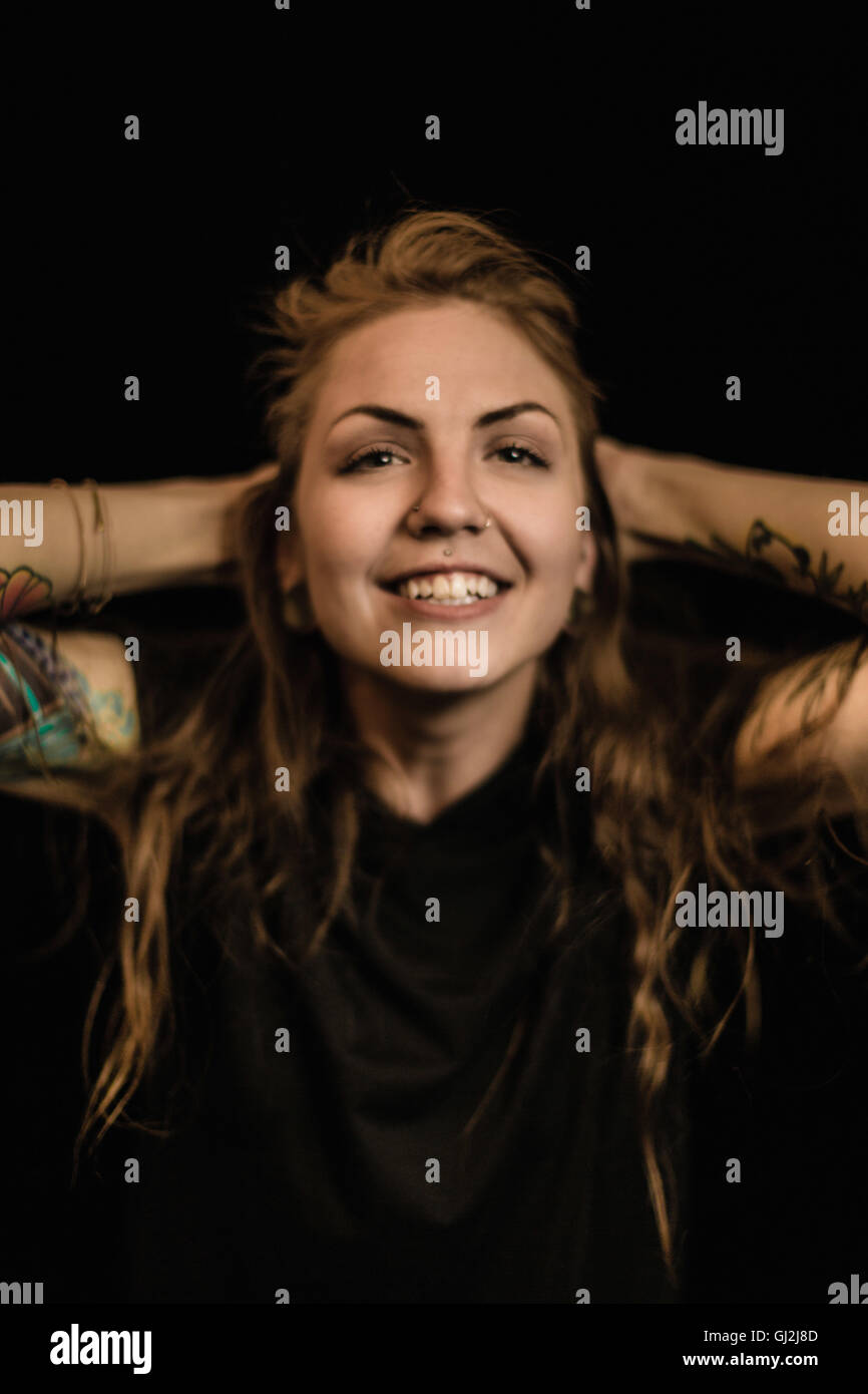 Portrait de femme avec des piercings et tatouages, les mains derrière la tête à la caméra en souriant Banque D'Images