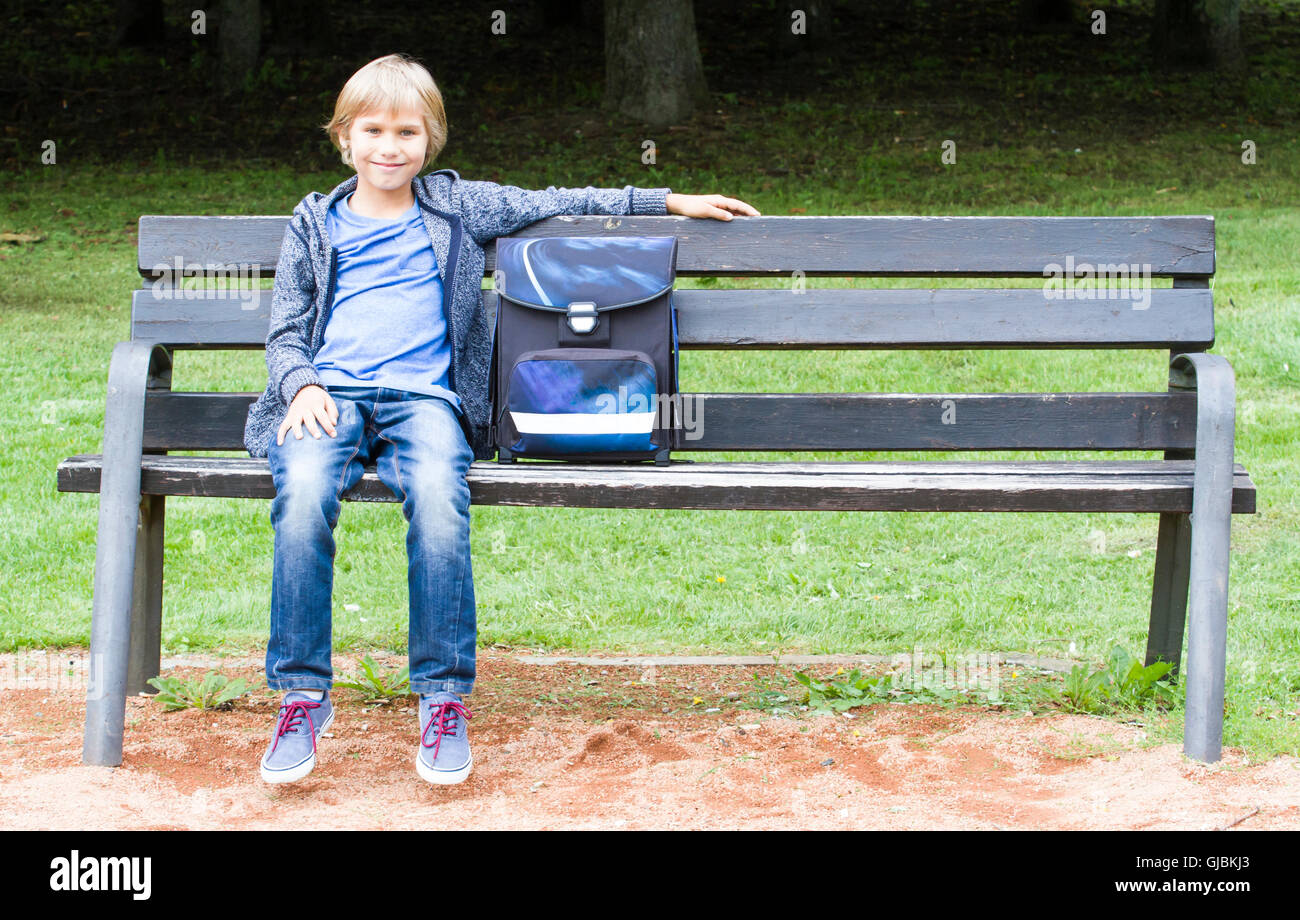 Smiling petit garçon assis sur un banc en bois avec son sac à dos. L'éducation, l'école, de vie, de personnes concept Banque D'Images