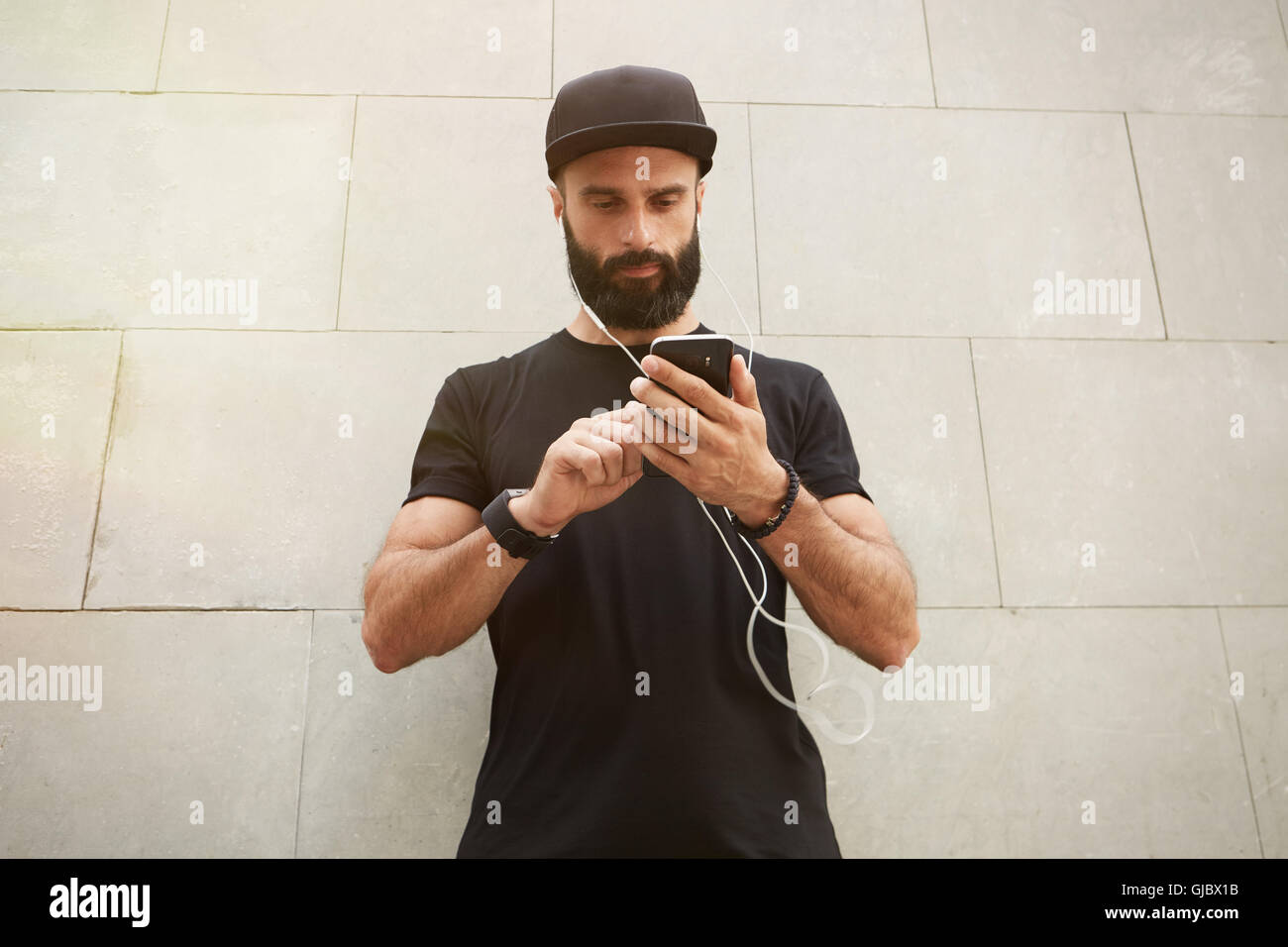 Homme musclé barbu vêtu de noir en blanc Tshirt Cap Snapback l'heure d'été.Les jeunes hommes debout en face du mur de béton gris vide arrière-plan à l'aide d'écouteurs Smartphone.Maquette horizontale. Banque D'Images