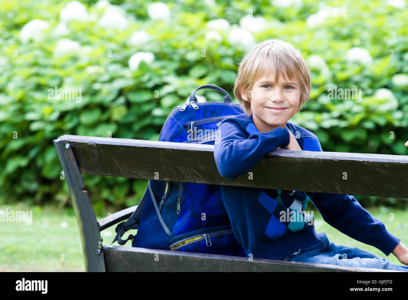 Smiling petit garçon assis sur un banc en bois avec son sac à dos. L'éducation, l'école, de vie, de personnes concept Banque D'Images