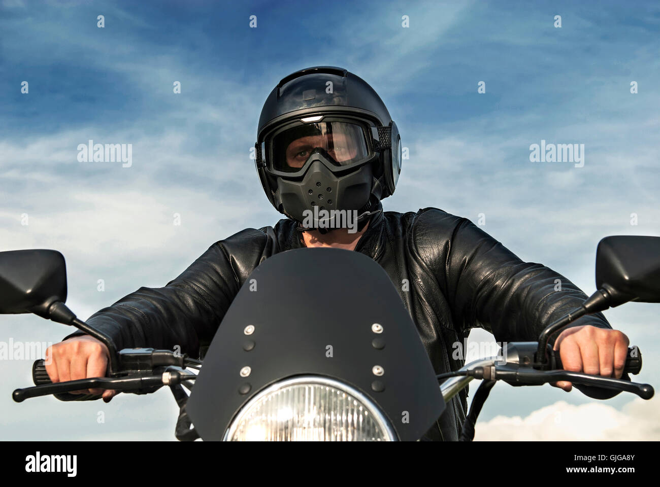 Motocycliste Motorbiker assis sur moto avec casque noir veste en cuir pare-soleil Ciel bleu nuages blancs les mains sur les poignées Banque D'Images