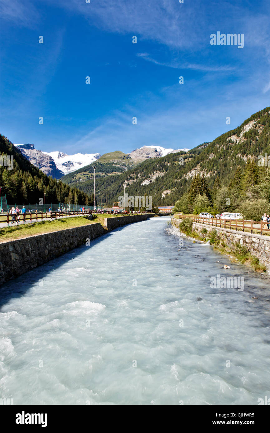 Rivière qui coule et des sommets alpins enneigés dans Champoluc en Italie du nord avec des gens qui marchent le long de la rivière vers lointain Banque D'Images