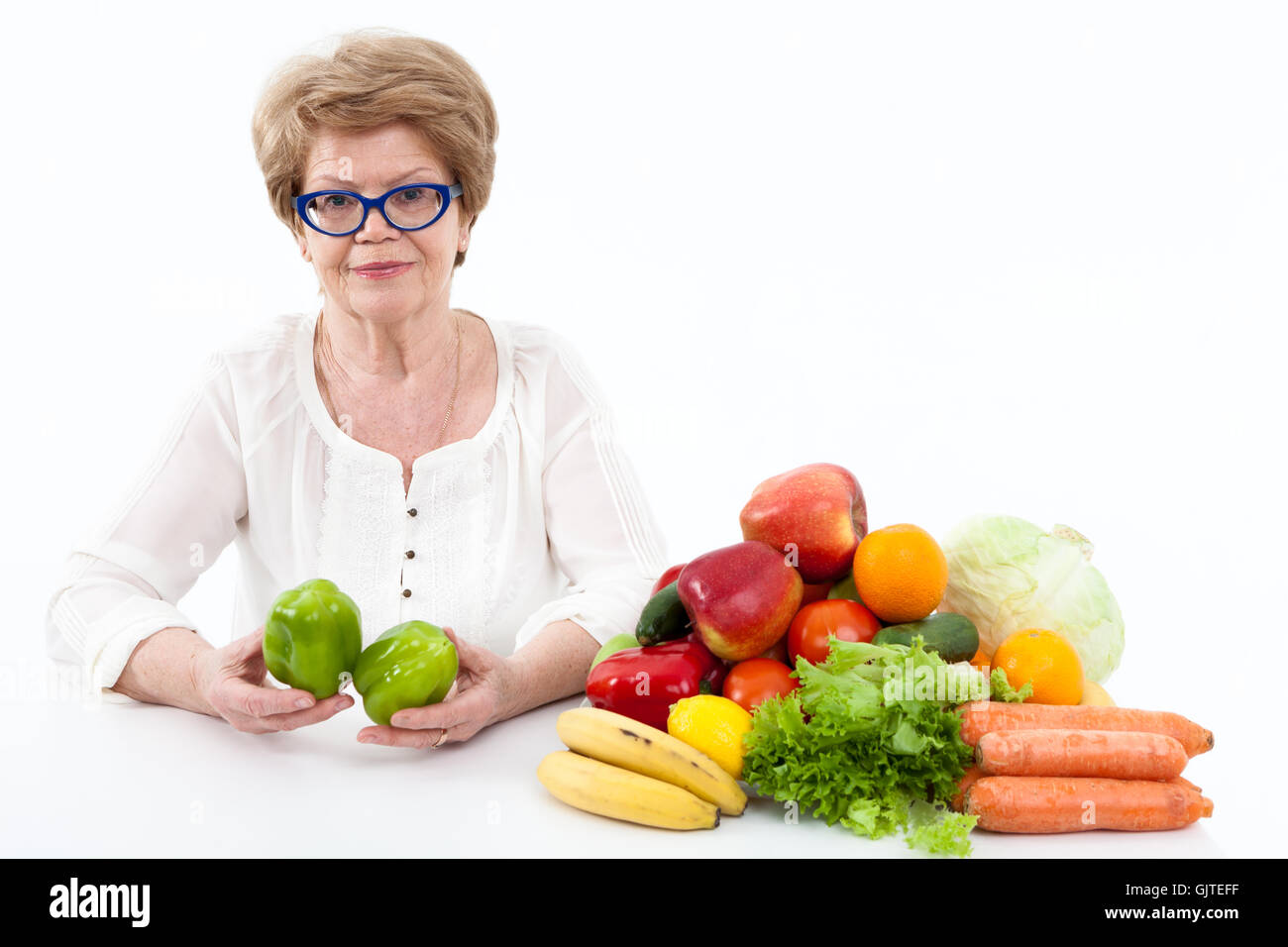 Aîné heureux Caucasian woman holding hands with deux poivrons verts, légumes et fruits sont sur la table, fond blanc Banque D'Images