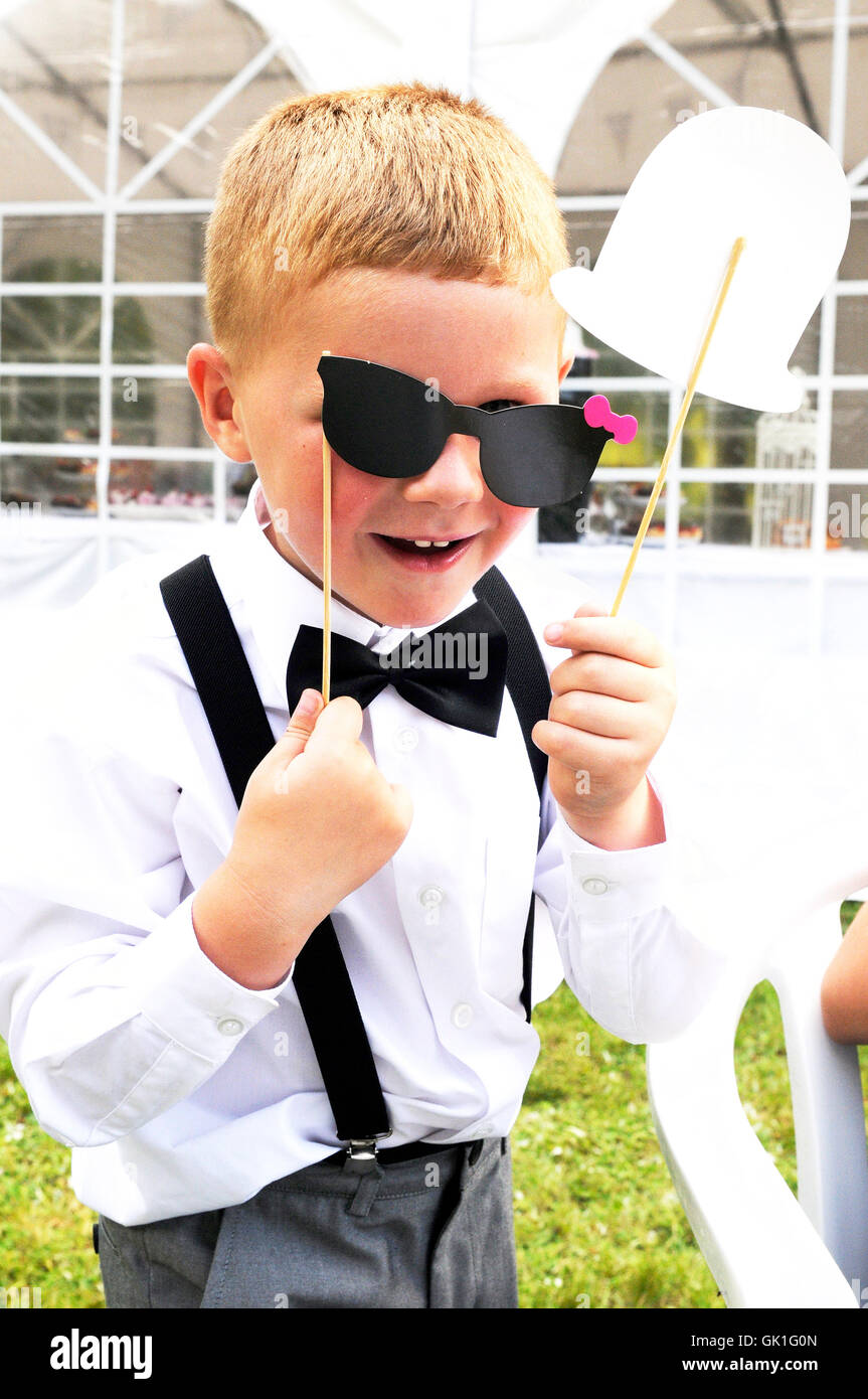 Jeune garçon à une réception de mariage,tenant une paire de lunettes en papier sur les yeux. Vêtu d'un costume du matin, avec papillon. Banque D'Images