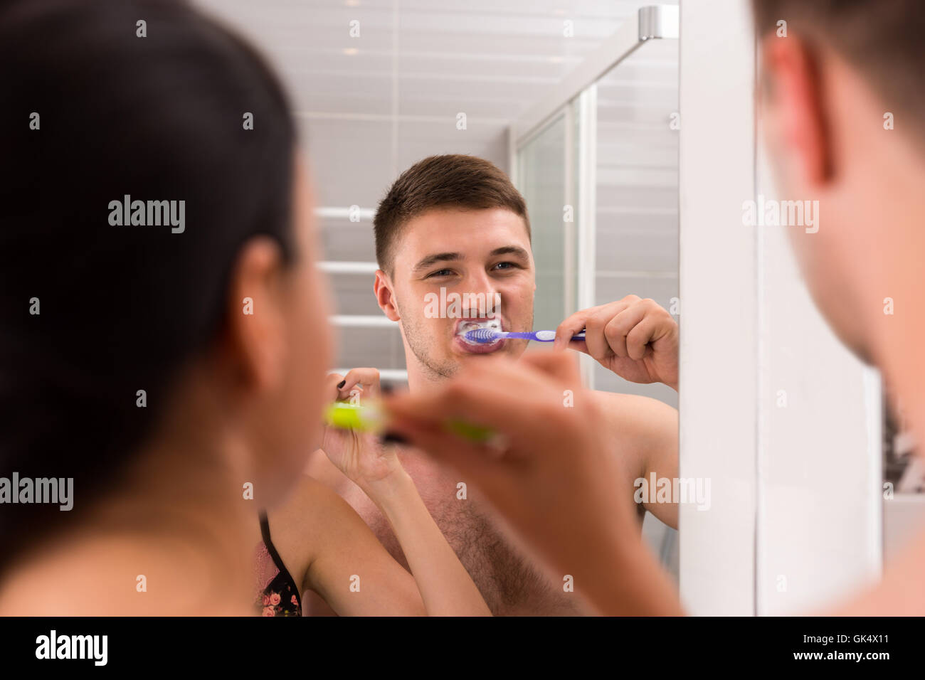 Portrait jeune homme se brosser les dents, debout devant un miroir de près de sa petite amie dans la salle de bains moderne carrelée à la maison Banque D'Images