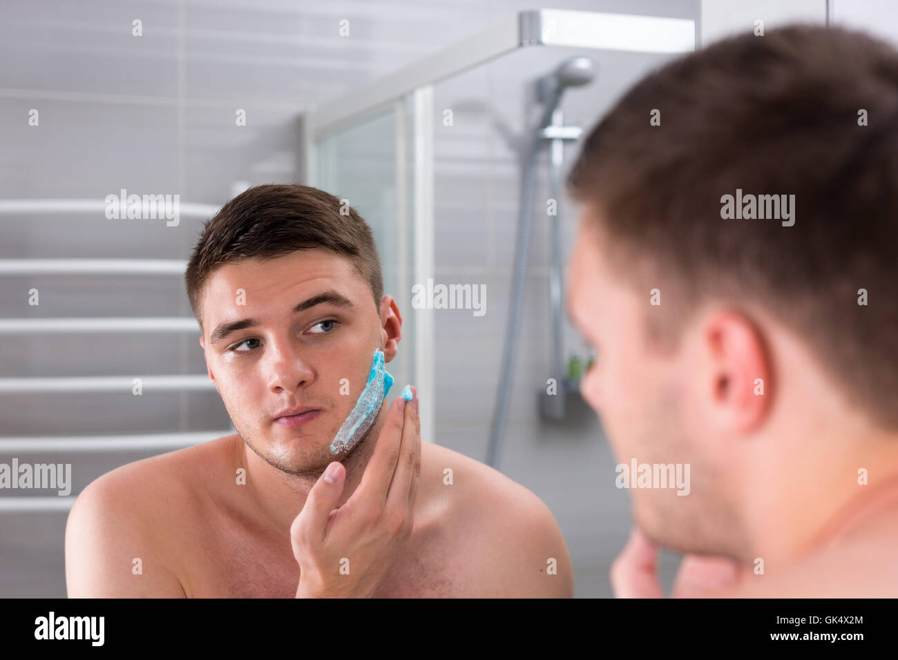 Beau jeune homme enduit de gel de rasage sur le visage en face de miroir dans la salle de bains moderne carrelée à la maison Banque D'Images