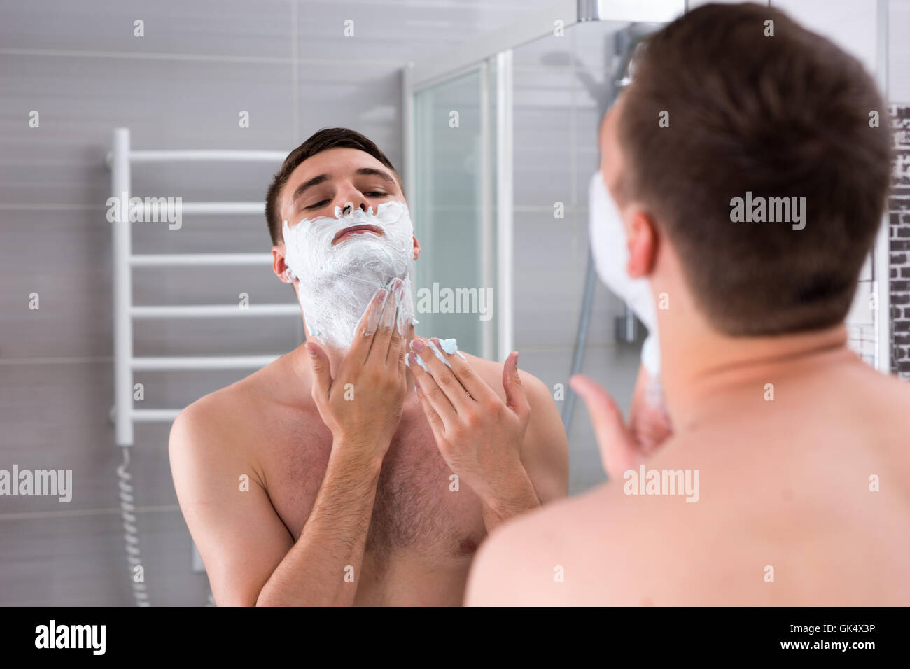 Jeune homme beau mâle pour gel de rasage shave sur son visage en face du miroir dans la salle de bains moderne carrelée à la maison Banque D'Images
