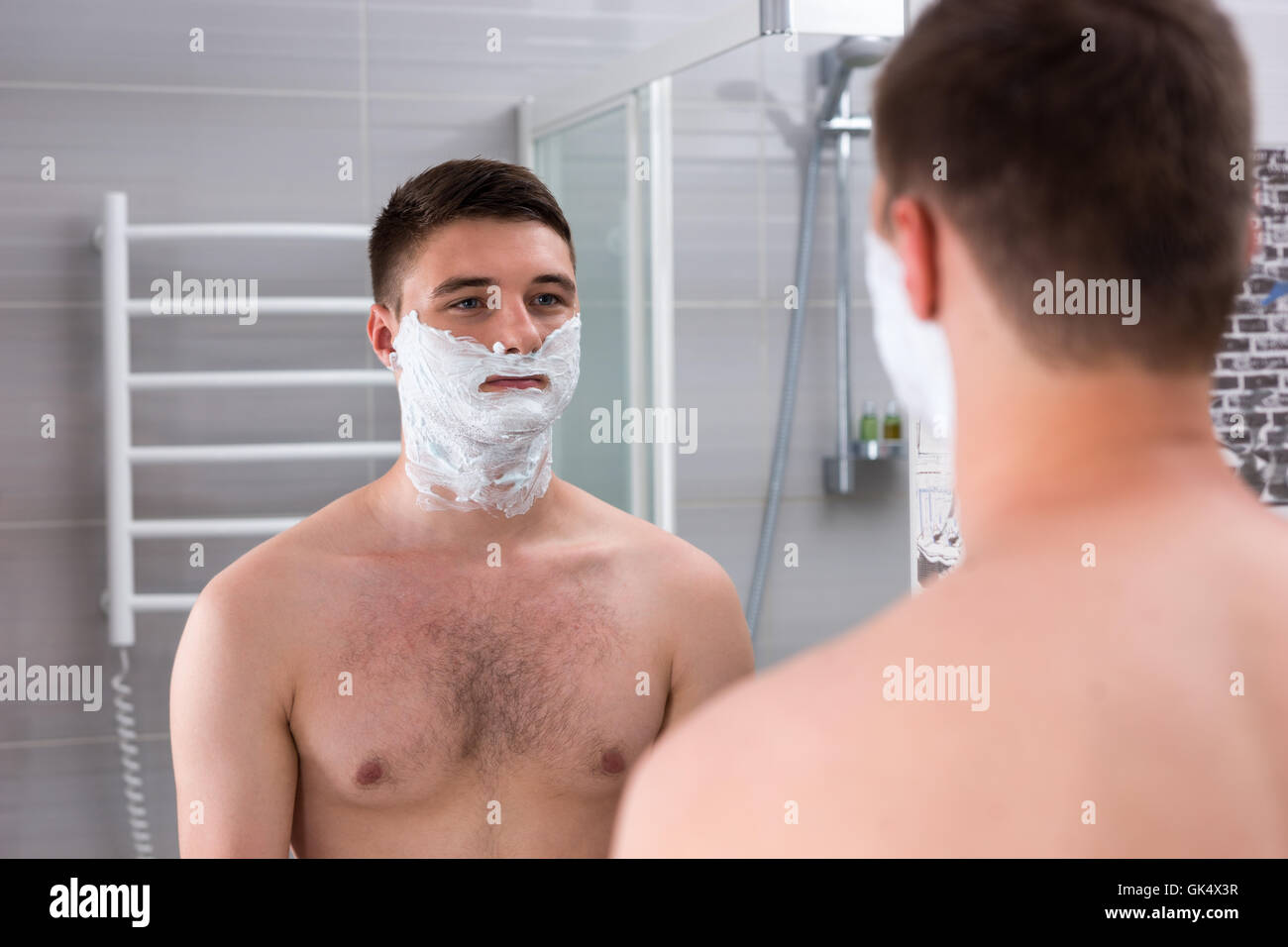 Joli jeune homme à la mousse à raser sur ses joues debout devant un miroir dans la salle de bains moderne carrelée à la maison Banque D'Images