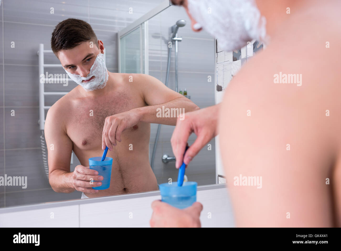 L'homme avec de la mousse sur ses joues se lave son rasoir dans la tasse avec de l'eau debout devant un miroir dans la salle de bains moderne carrelée à la maison. Banque D'Images