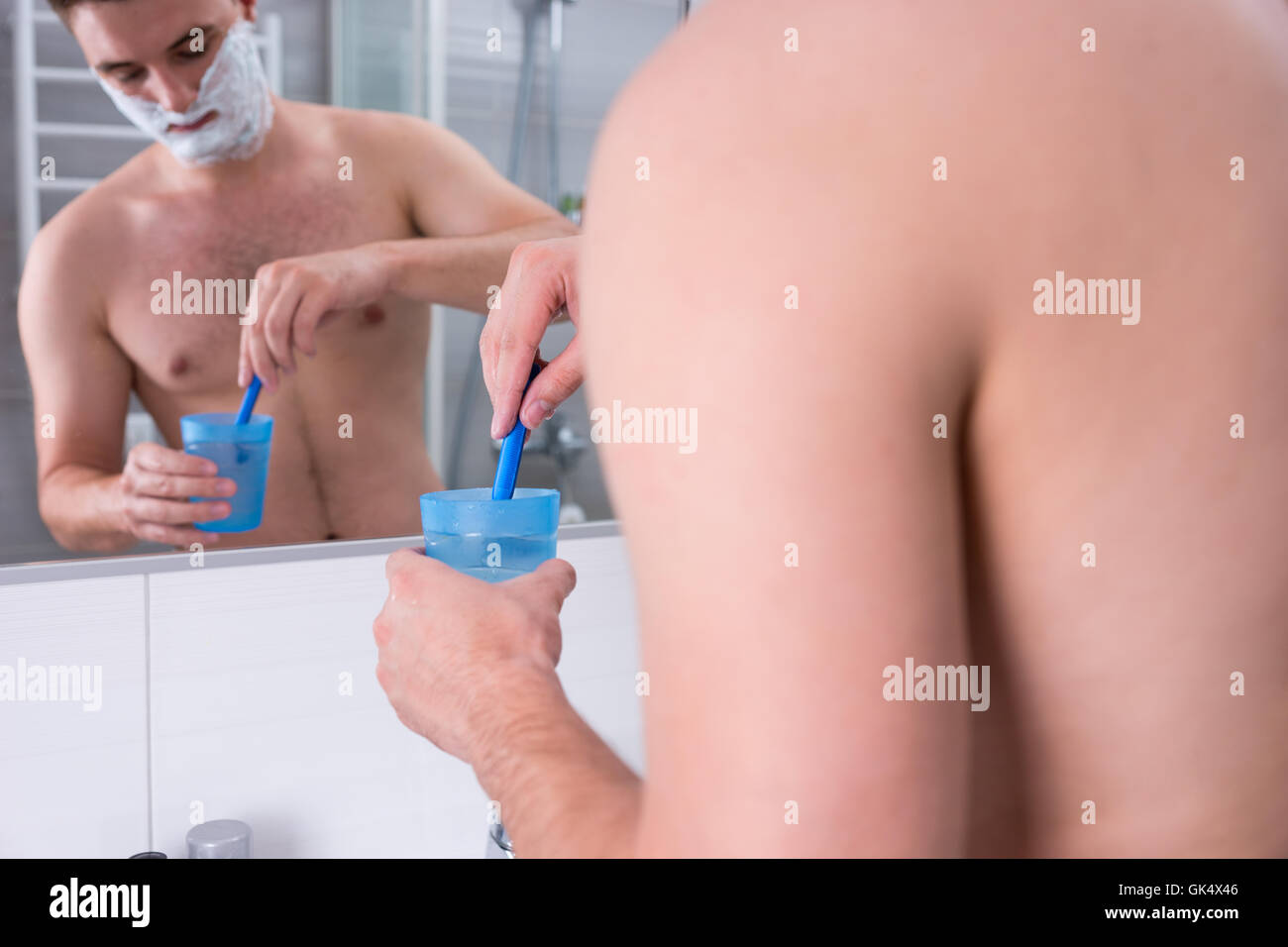 Jeune homme à la mousse à raser sur ses joues se lave son rasoir dans la tasse avec de l'eau debout devant un miroir dans la salle de bains moderne carrelée. Banque D'Images