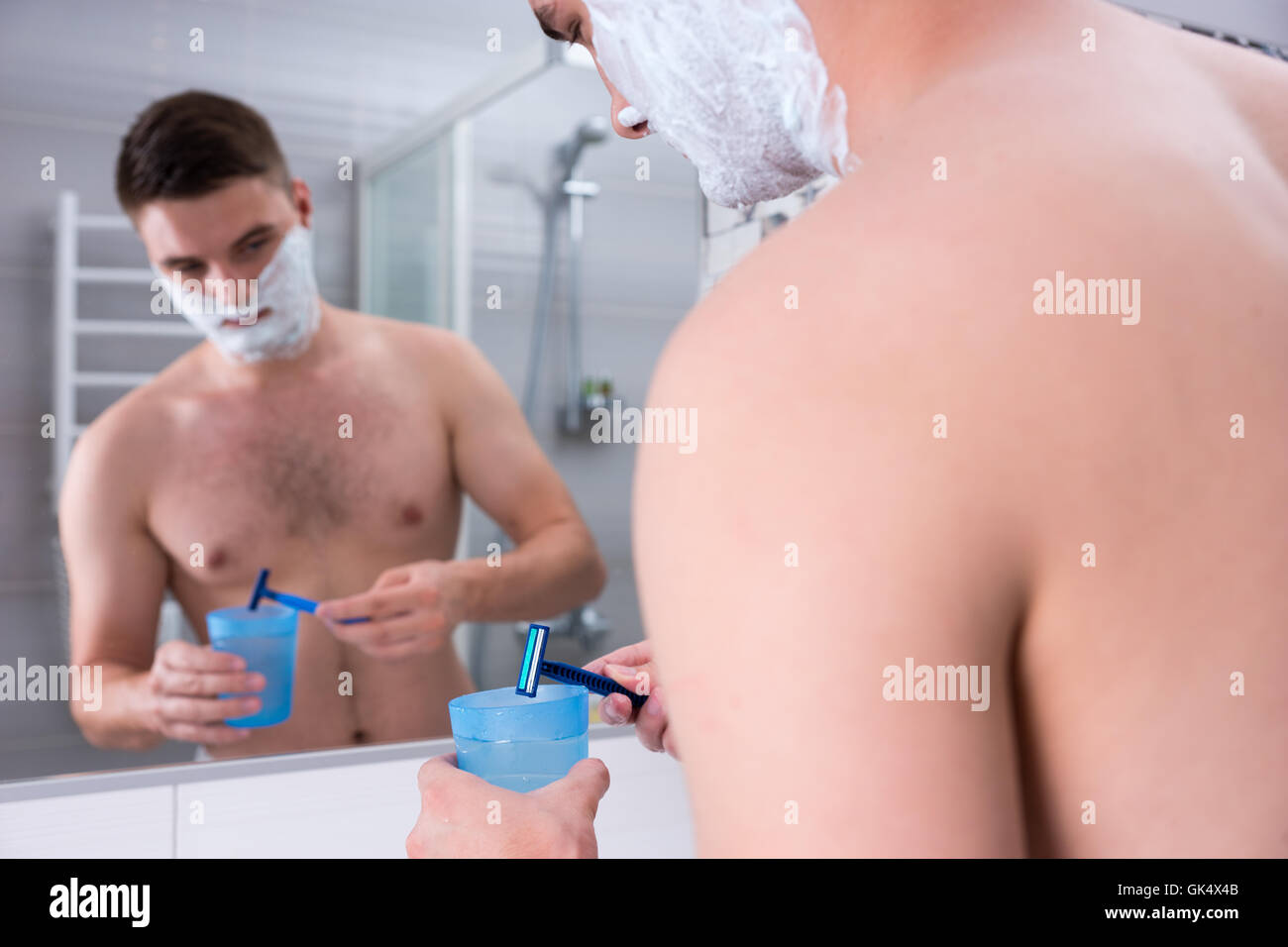 Beau mâle avec de la mousse sur ses joues se lave son rasoir dans la tasse avec de l'eau debout devant un miroir dans la salle de bains moderne carrelée à la maison. Banque D'Images