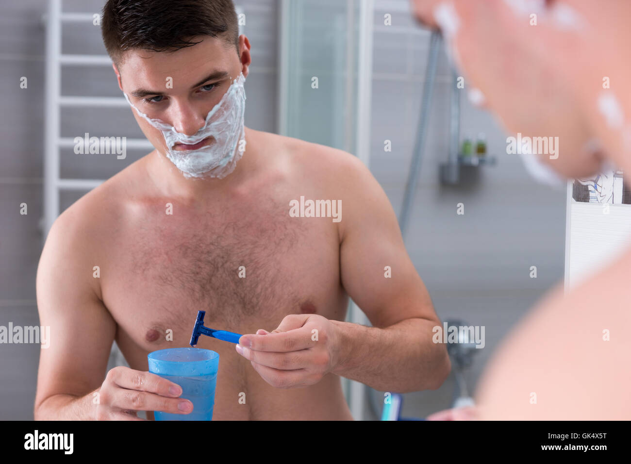Man un rasoir dans la tasse avec de l'eau debout devant un miroir dans la salle de bains moderne carrelée à la maison Banque D'Images