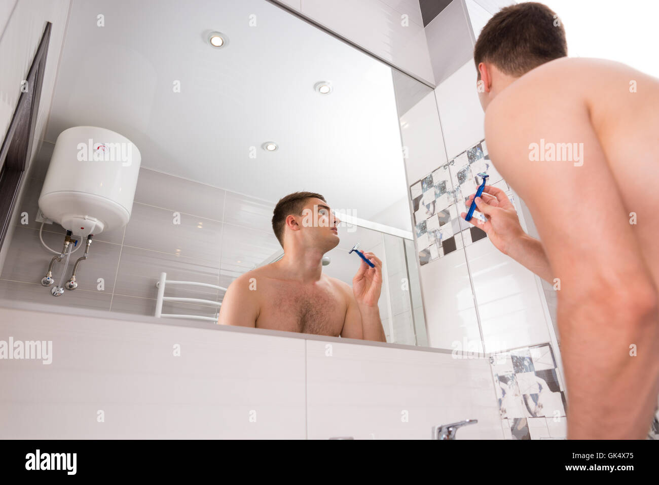 Vue de dessous du jeune homme se raser le visage avec un razer debout devant un miroir dans la salle de bains moderne carrelée à la maison Banque D'Images