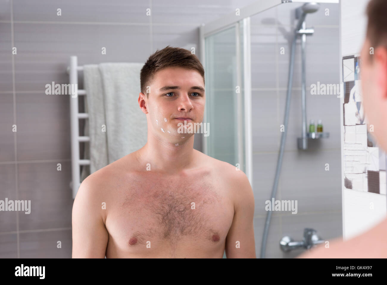 Jeune homme avec des coupures sur son visage après rasage mauvais debout devant un miroir dans la salle de bains moderne carrelée à la maison Banque D'Images