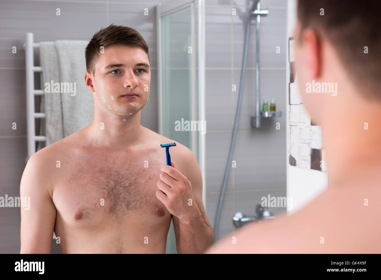 Jeune homme avec des coupures sur son visage après rasage mauvais holding un rasoir alors qu'il se tenait devant le miroir de la salle de bains moderne carrelée à la maison. Banque D'Images