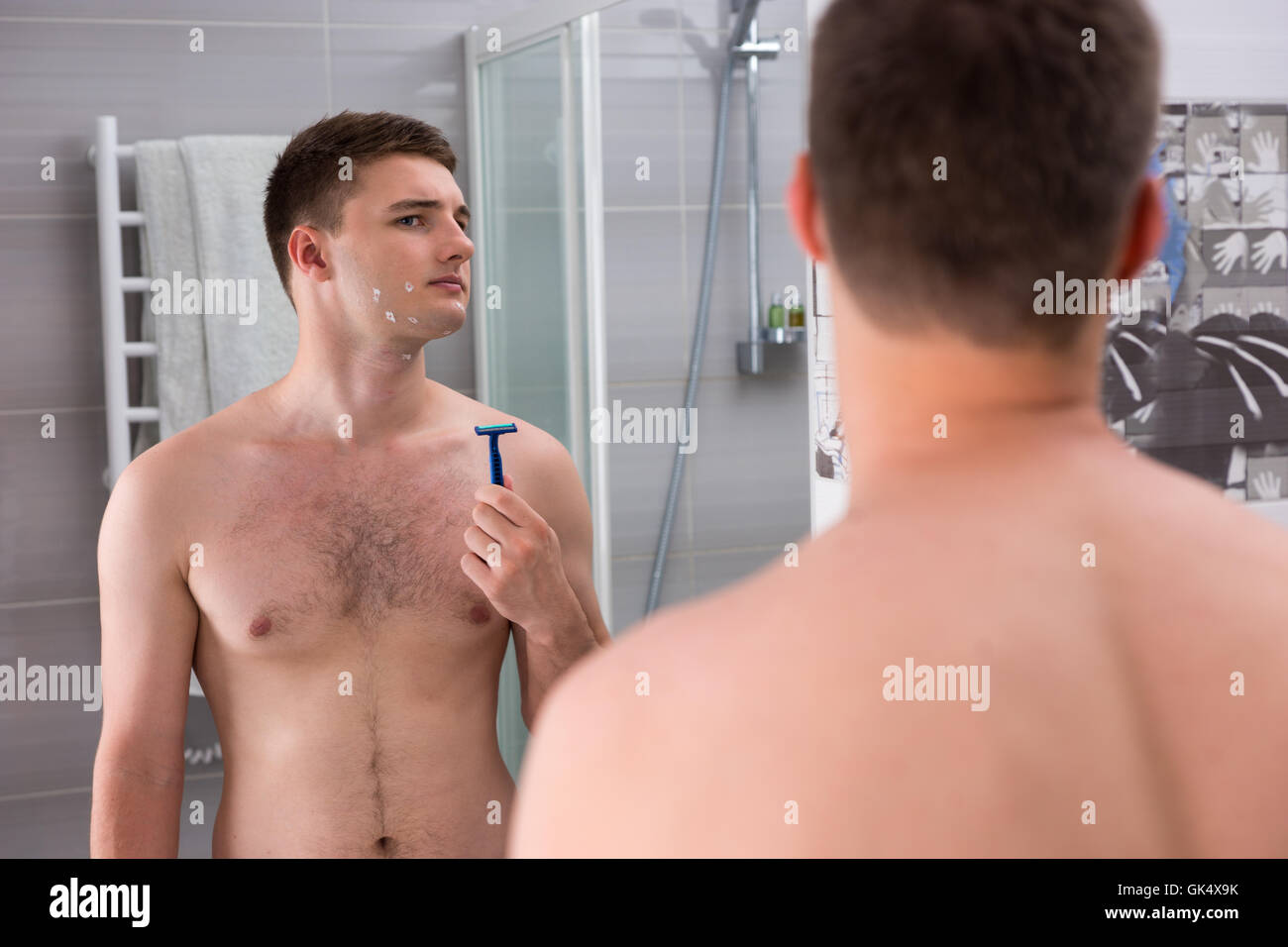 Homme avec des coupures sur son visage après rasage mauvais holding un rasoir alors qu'il se tenait devant le miroir de la salle de bains moderne carrelée à la maison. Banque D'Images