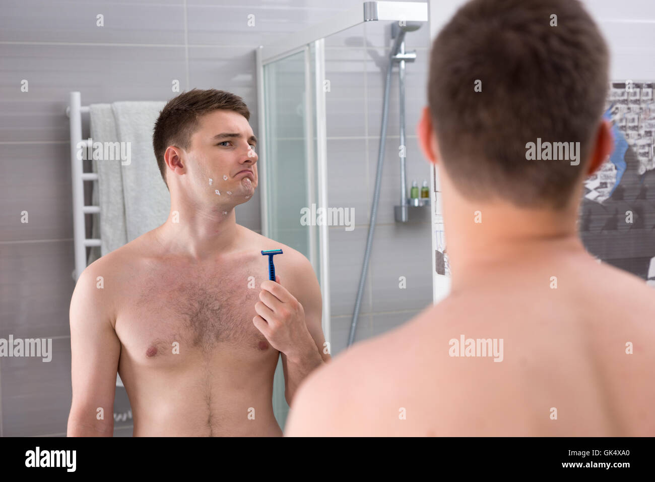 Jeune homme mécontent avec des coupures sur son visage après rasage mauvais holding un rasoir alors qu'il se tenait devant le miroir de la salle de bains moderne carrelée à la maison. Banque D'Images