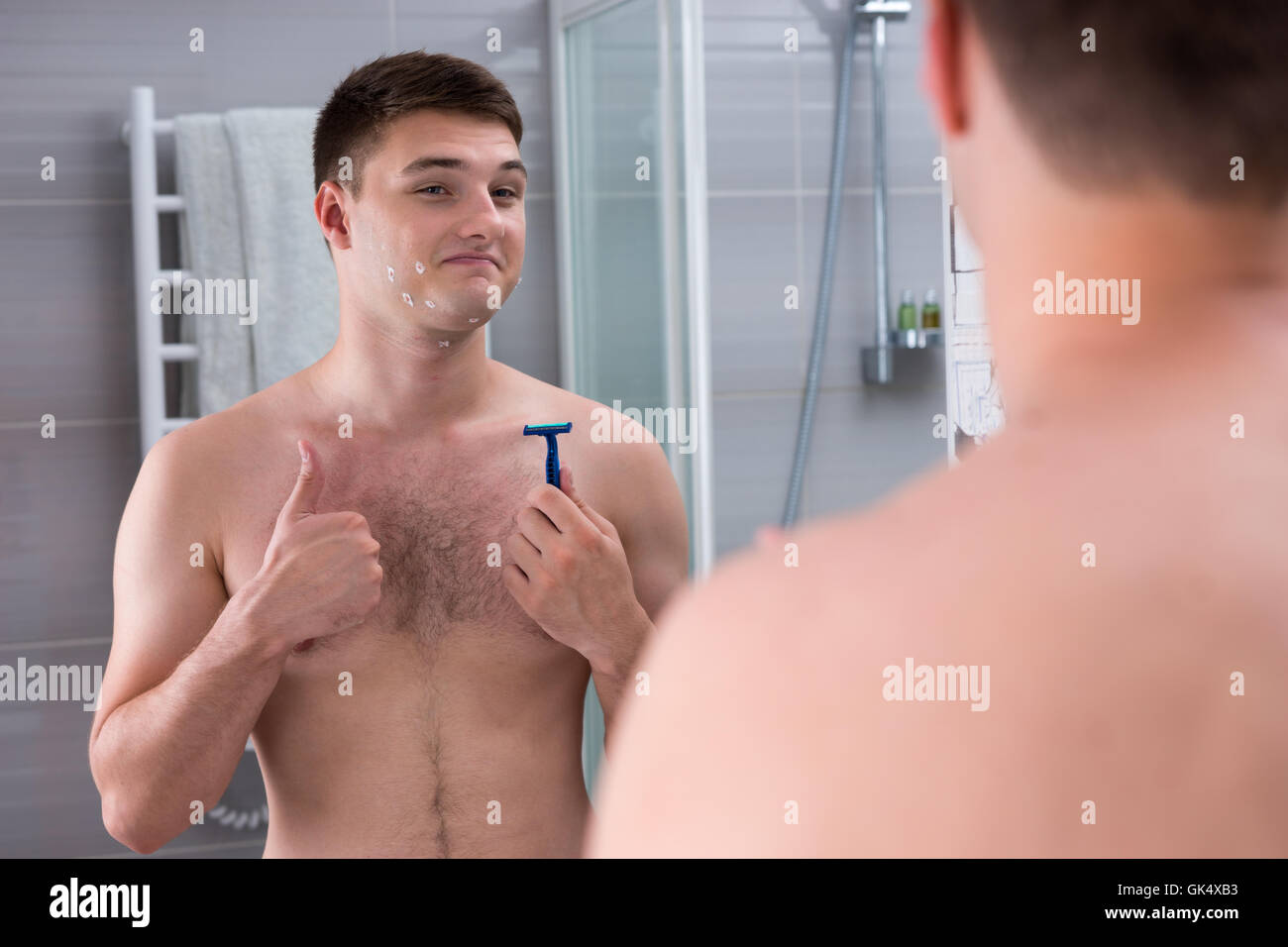 Heureux jeune homme avec des coupures sur son visage maintenant un rasoir alors qu'il se tenait devant le miroir de la salle de bains moderne carrelée à la maison. Banque D'Images