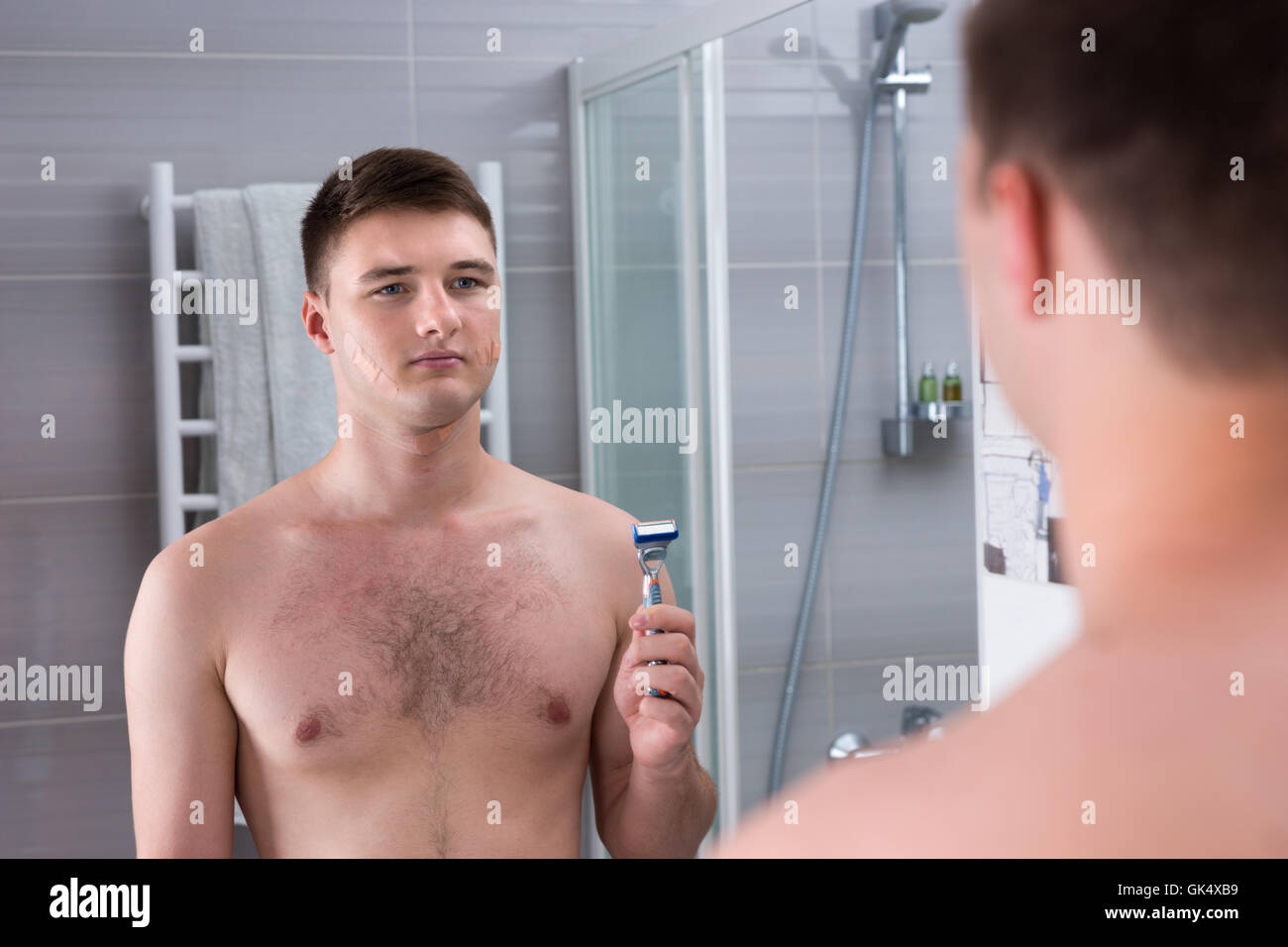 Après rasage homme mauvais avec des cataplasmes sur son visage maintenant  un rasoir alors qu'il se tenait devant le miroir de la salle de bains  moderne carrelée à la maison Photo Stock -