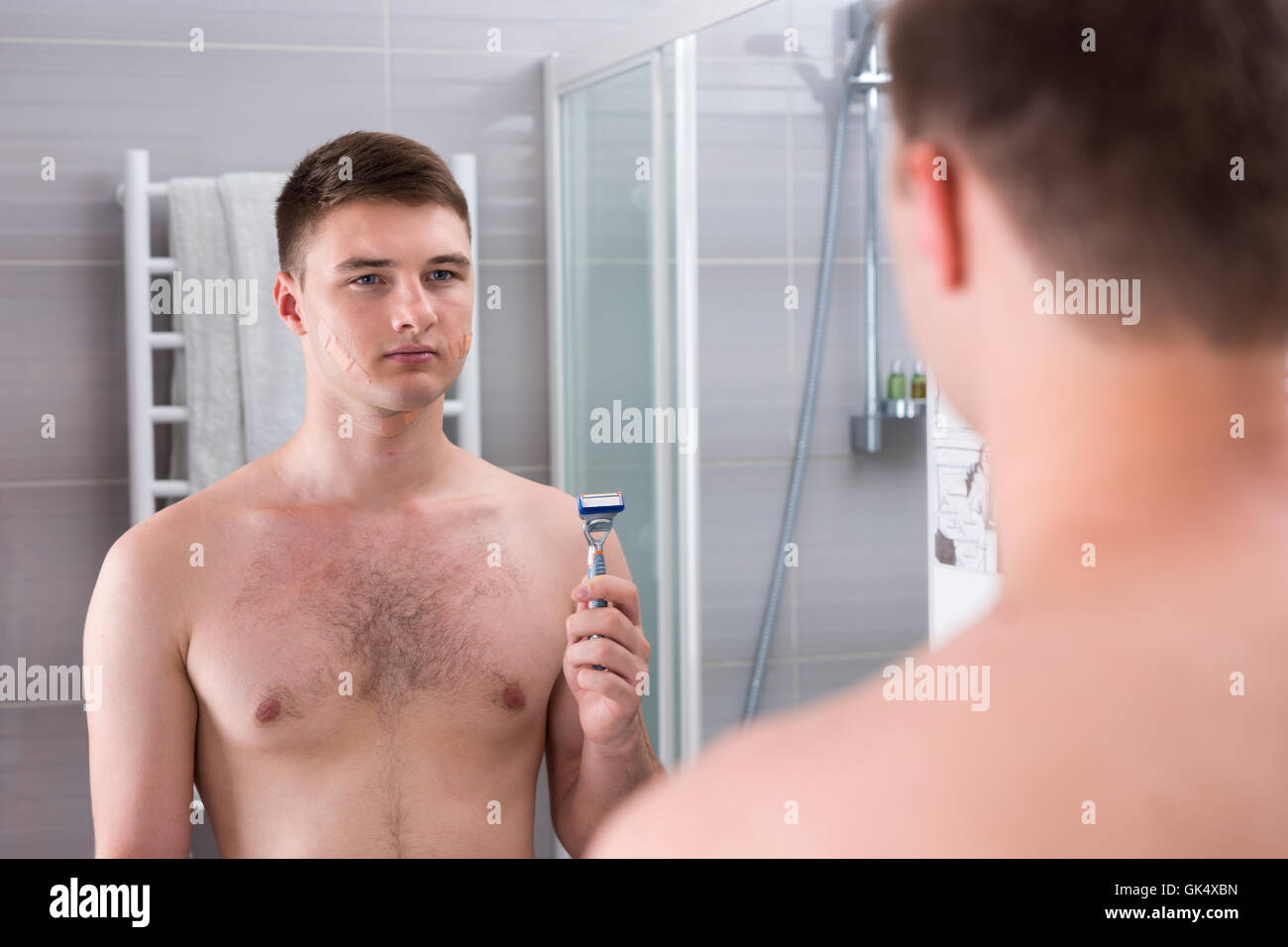 Beau jeune homme après rasage mauvais avec des cataplasmes sur son visage maintenant un rasoir alors qu'il se tenait devant le miroir de la salle de bains moderne carrelée à la maison. Banque D'Images