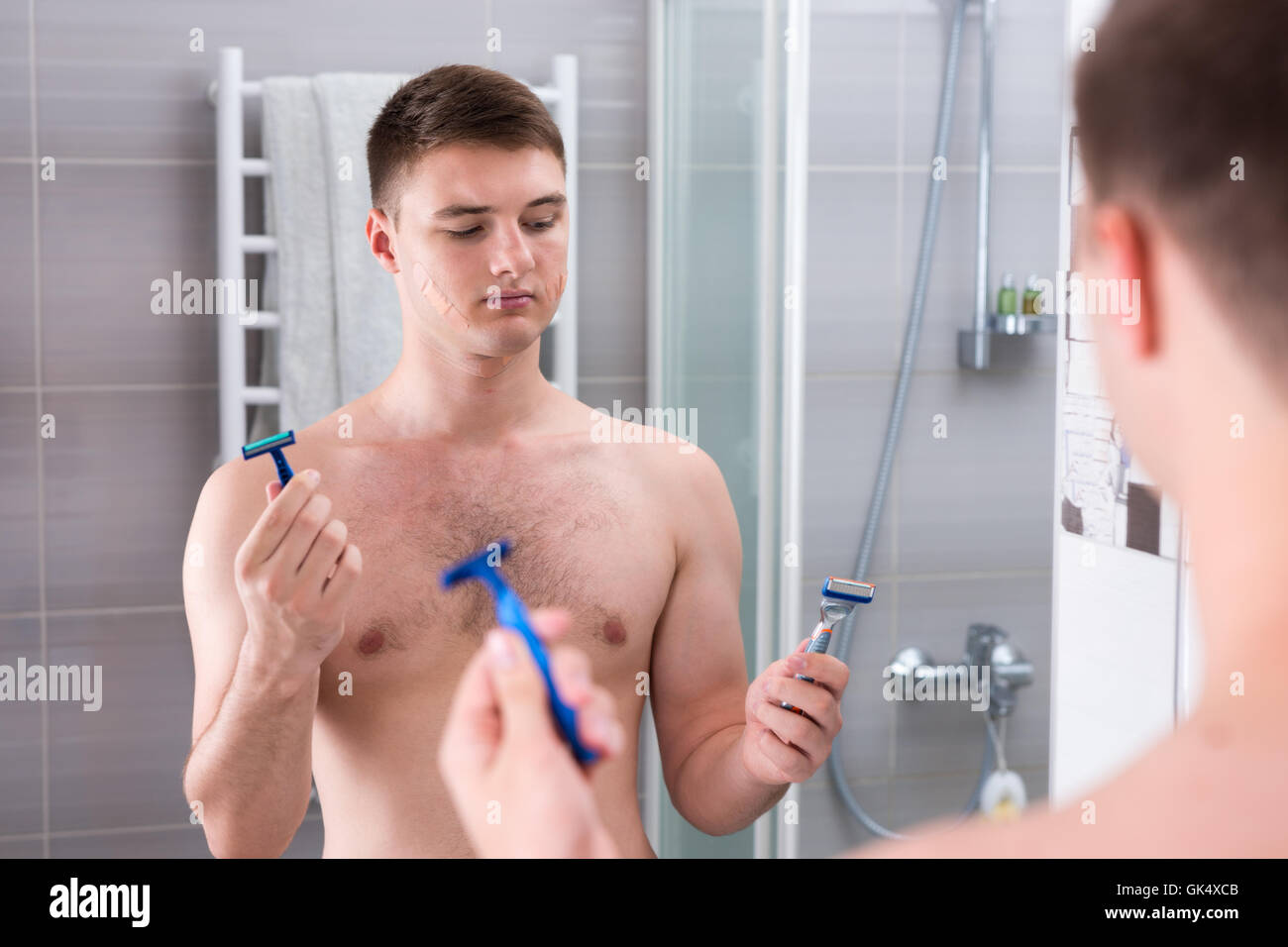 Jeune homme tenant les rasoirs et choisit le meilleur alors qu'il se tenait devant le miroir de la salle de bains moderne carrelée à la maison Banque D'Images