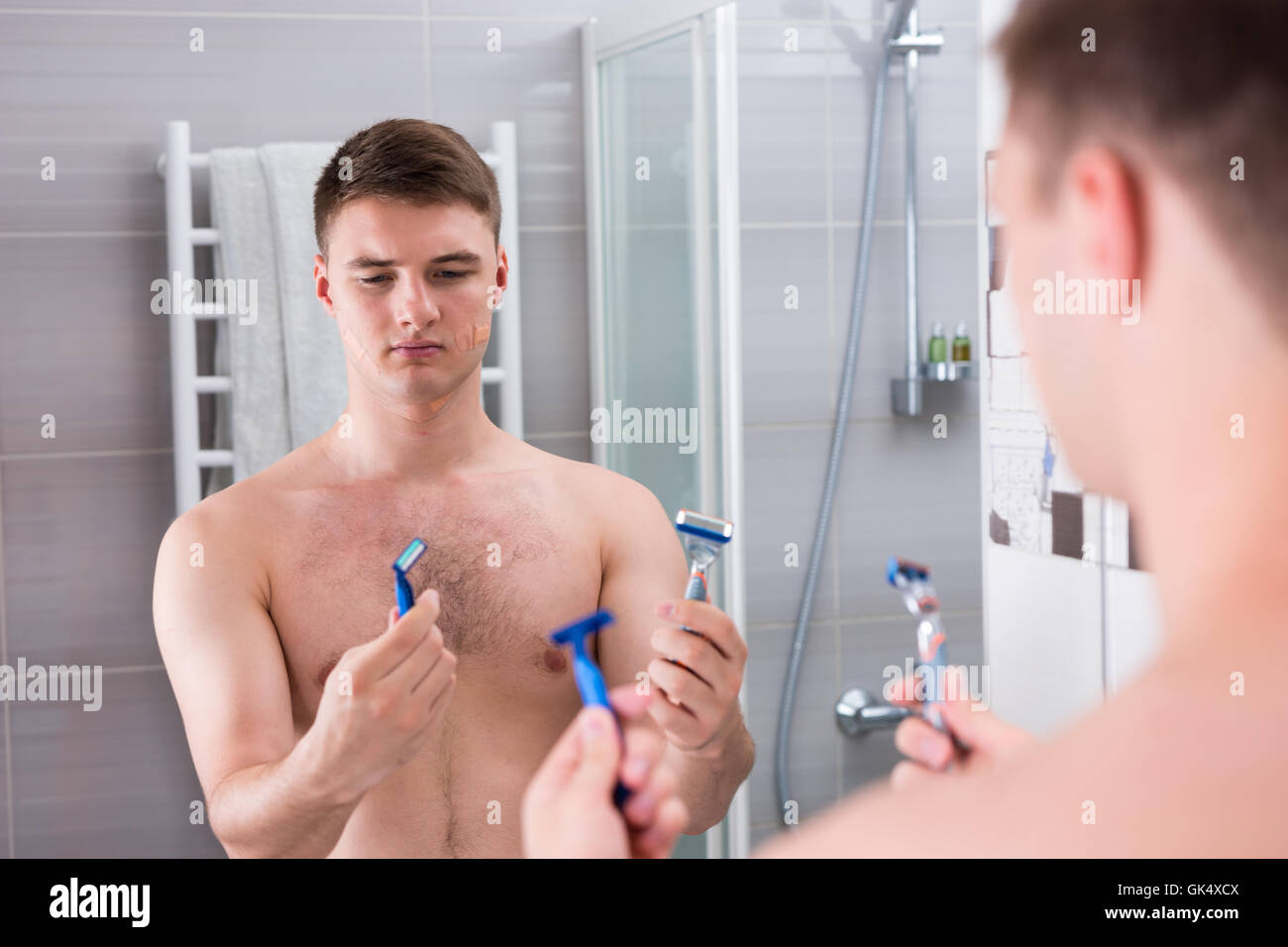 Handsome man holding rasoirs et choisit le meilleur alors qu'il se tenait devant le miroir de la salle de bains moderne carrelée à la maison Banque D'Images