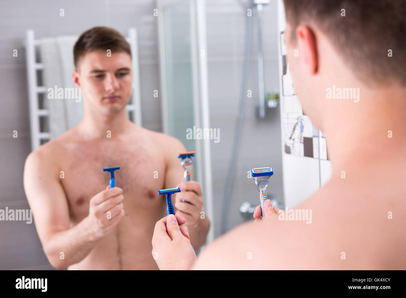 Les rasoirs masculins holding et choisit le meilleur alors qu'il se tenait devant le miroir de la salle de bains moderne carrelée à la maison Banque D'Images