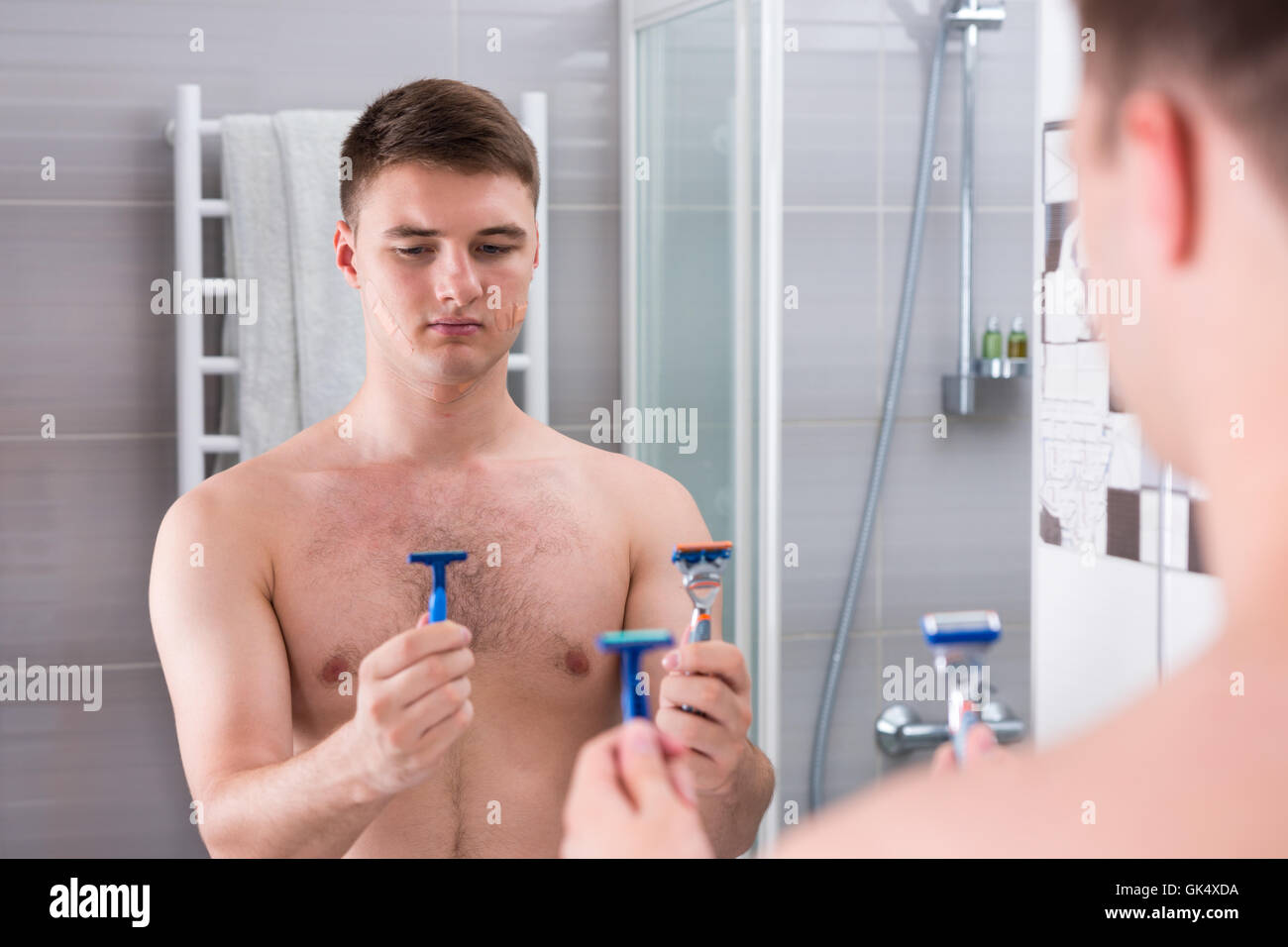 Beau Jeune homme tenant les rasoirs et choisit le meilleur alors qu'il se tenait devant le miroir de la salle de bains moderne carrelée à la maison. Banque D'Images