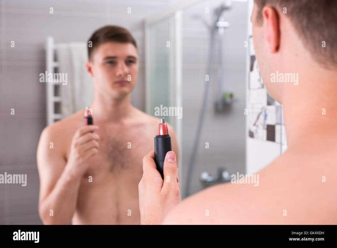 Jeune homme tenant une tondeuse alors qu'il se tenait devant le miroir de la salle de bains moderne carrelée à la maison Banque D'Images