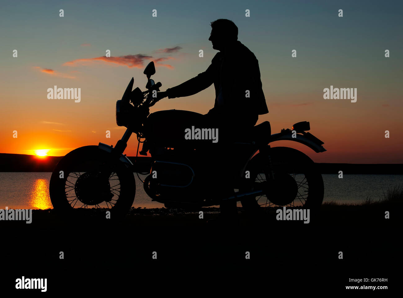 Motorbiker motocycliste en silhouette au coucher du soleil par le lac de l'homme en moto une main au guidon Banque D'Images