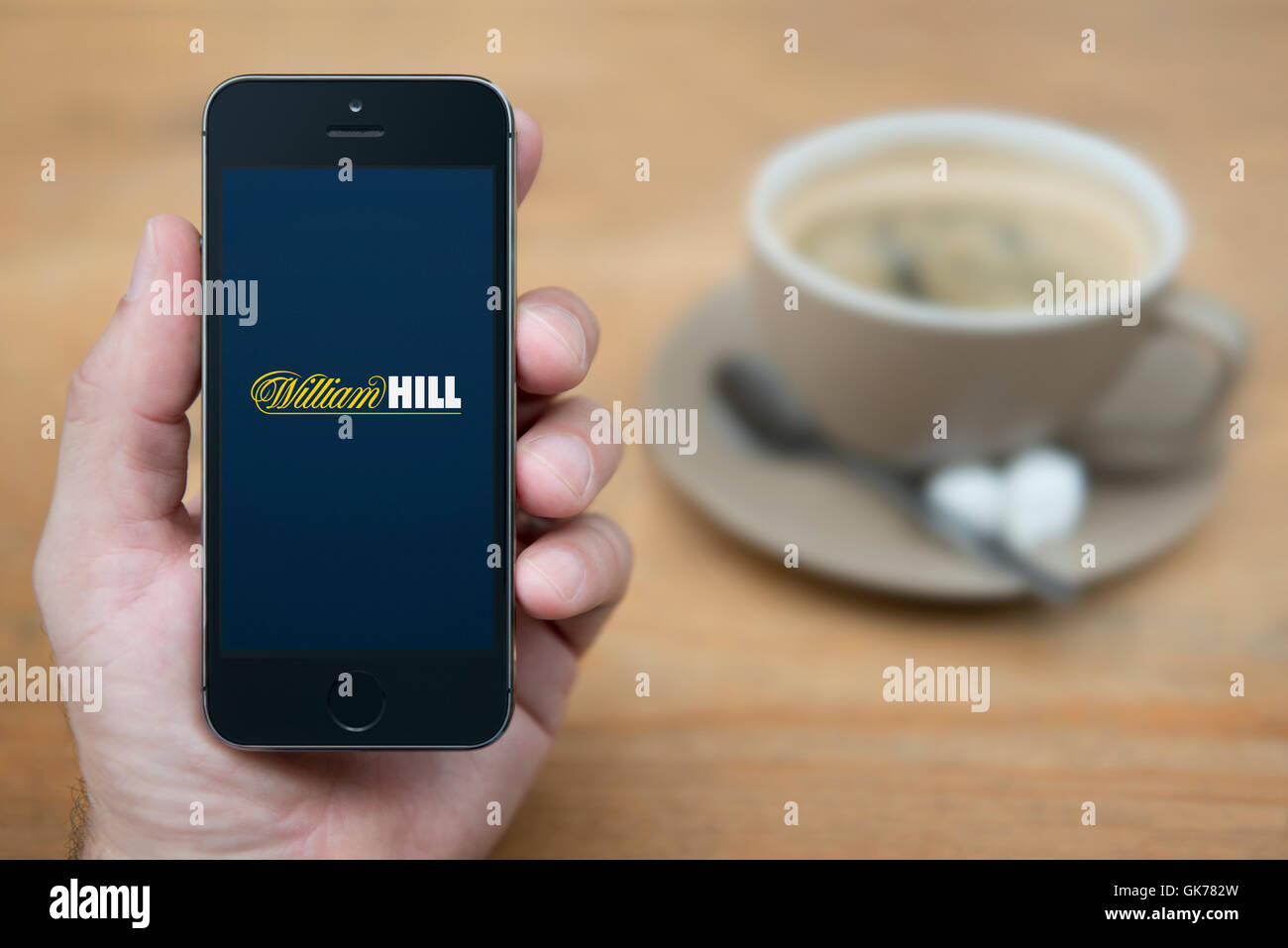 Un homme se penche sur son iPhone qui affiche le logo de William Hill, alors qu'assis avec une tasse de café (usage éditorial uniquement). Banque D'Images