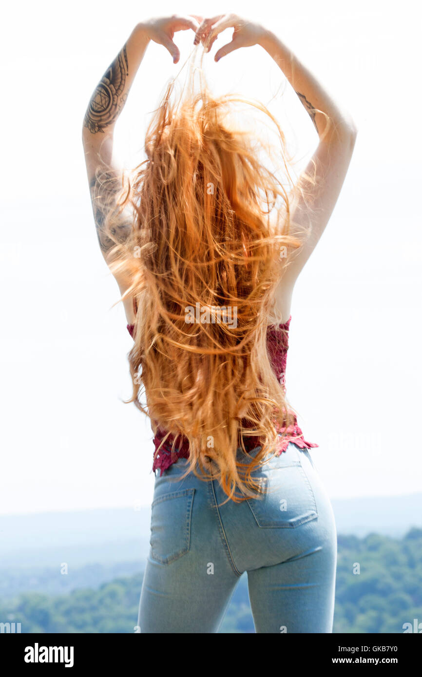 Vue arrière de belle tête rouge en skinny jeans et haut rouge, en montagne au soleil d'or, le vent dans les cheveux et les bras levés. Banque D'Images
