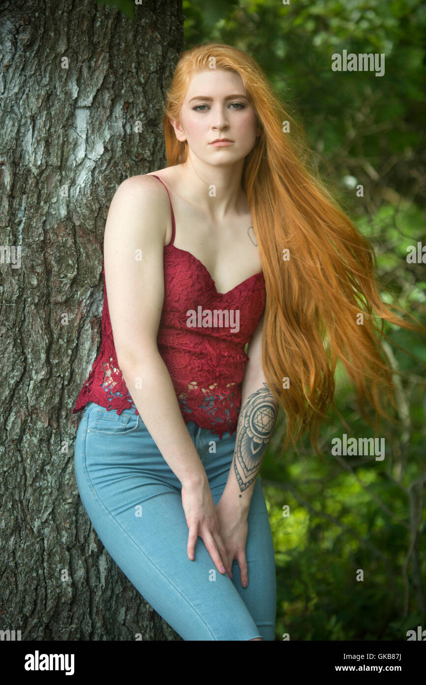Belle tête rouge femme en skinny jeans et haut rouge, se penchant presque pleine longueur contre un arbre. Banque D'Images